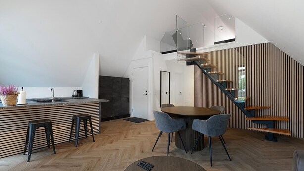 A modern kitchen with a central island, a dining area with a round table and chairs, and a staircase with a glass railing.
