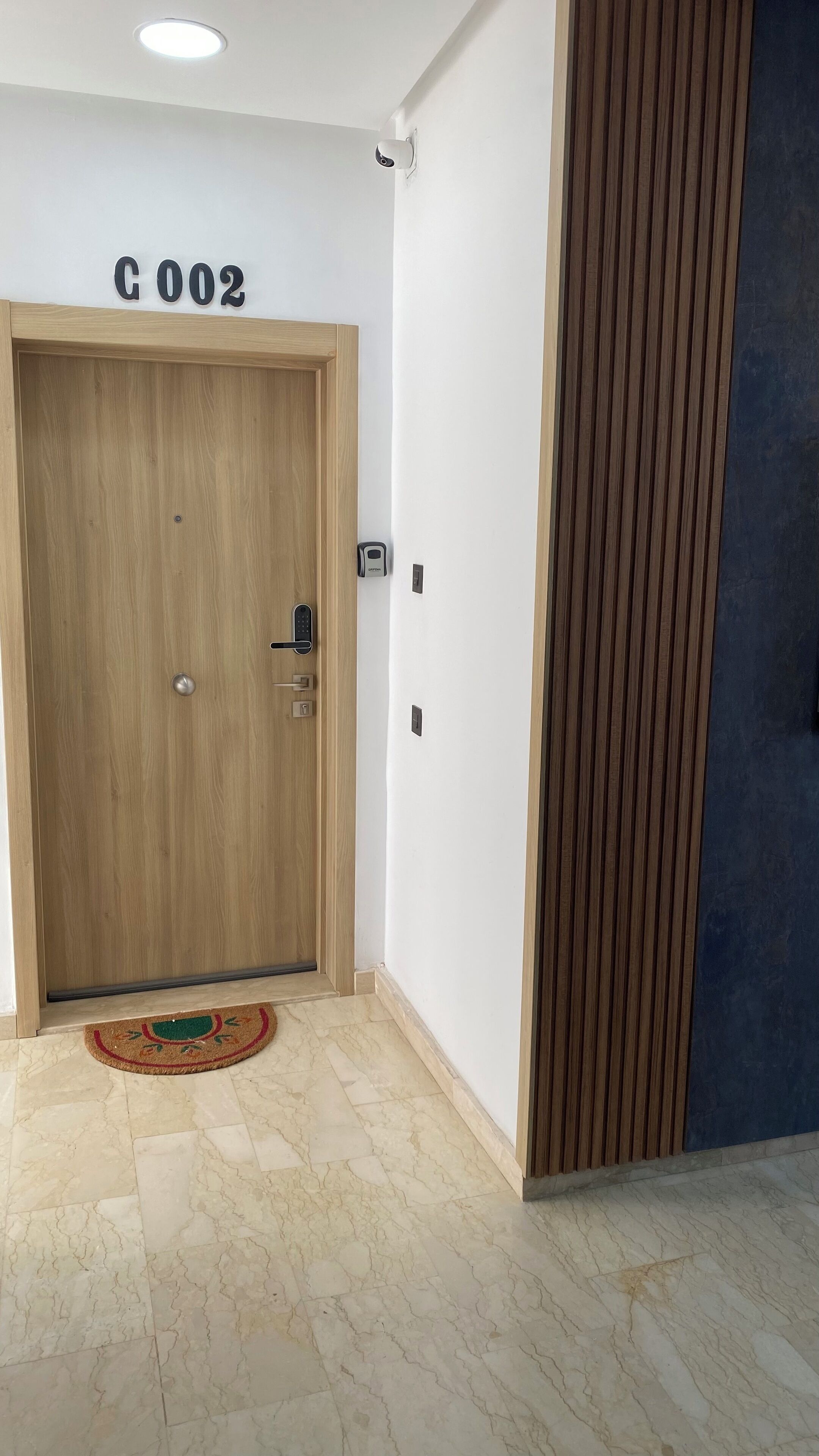 A wooden door with a keypad lock, a security camera above it, and a mat at the entrance.