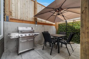 Une terrasse extérieure équipée d’un barbecue en acier inoxydable, d’une table, de chaises et d’un grand parasol.