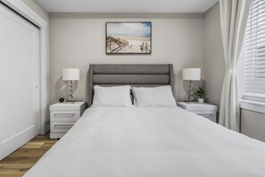 Une chambre à coucher avec un lit, deux tables de chevet, une petite table, une lampe fixée au mur, une plante en pot et un tableau représentant une scène de plage.