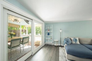 A room with a sliding glass door leading to an outdoor area, a sofa with a blue blanket, a white shelf unit, and a small round table with a lamp. - Island Time Escape - Charming Family Retreat (Key West)