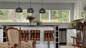 A modern kitchen with a central island, pendant lights, a dining area with striped chairs, and a view of greenery outside. - Entire Peaceful 3BR Guest Suite Upper Level West Vancouver: WiFi & Parking (West Vancouver)