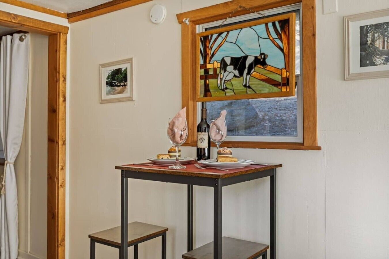 A room with a table set for two, a wine bottle, and a glass. There is a window with a stained glass depicting a cow.