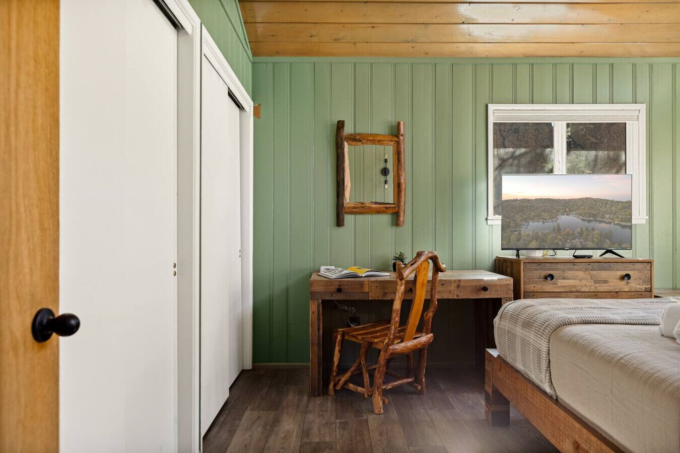 A bedroom with a wooden desk, a chair, a bed, a dresser, and a window with a view.