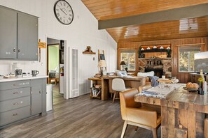 A rustic interior with a wooden ceiling, a kitchen area with grey cabinets, a dining table with chairs, and a stone fireplace. - Lark Lookout: Serene Surroundings on Huge Private Lot! Hot Tub & Walk to Lake! (Big Bear Lake)