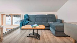 A modern living room with a grey sectional sofa, a wooden coffee table, and a view of a snowy landscape through a large window. - Chalet Arpitan Orizon - Chalet in the Vanoise National Park - OVO Network (Pralognan-la-Vanoise)