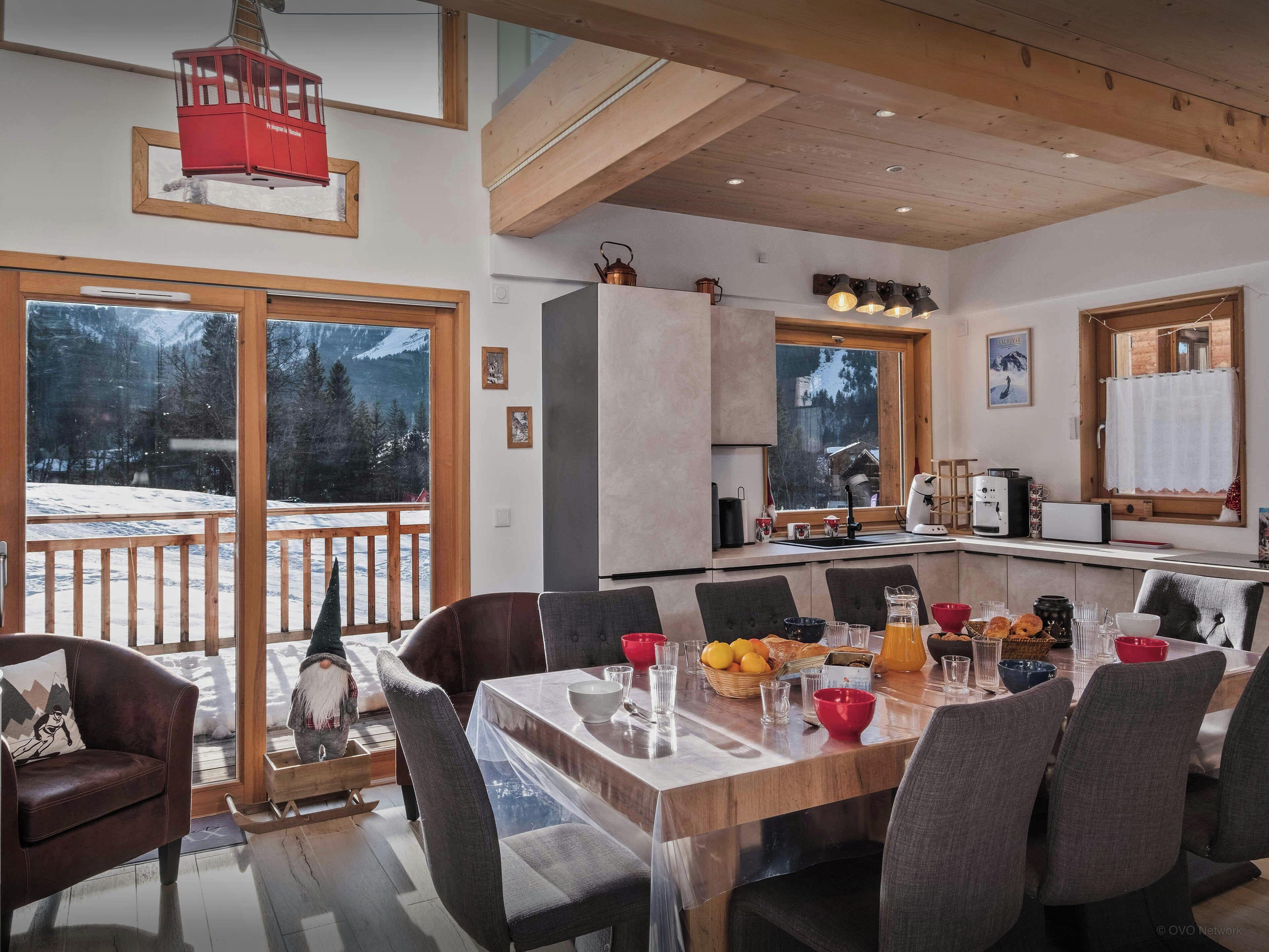 A modern kitchen with a large island, wooden ceiling beams, and large windows overlooking a snowy landscape.