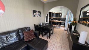 Un salon moderne avec un canapé en cuir noir, une table à manger avec des chaises, une cuisine en arrière-plan et un grand miroir sur le mur.