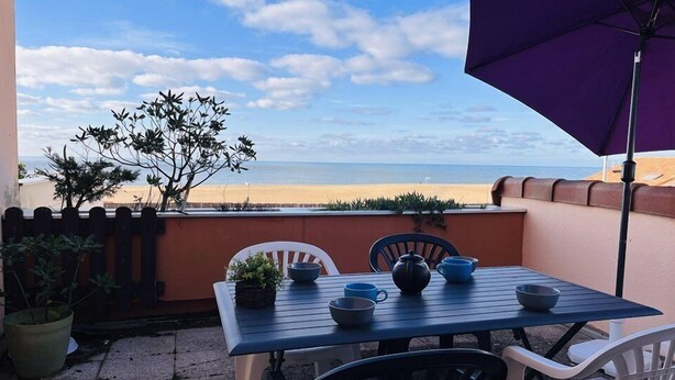 Ein Tisch mit Stühlen und einem violetten Sonnenschirm auf einem Balkon mit Blick auf einen Strand.