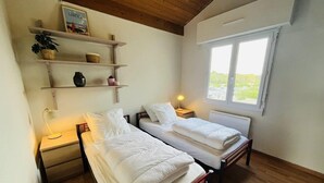 Ein Zimmer mit zwei Betten, Holzboden, einem Fenster, einem Regal mit Dekoration und einer Nachttischlampe.