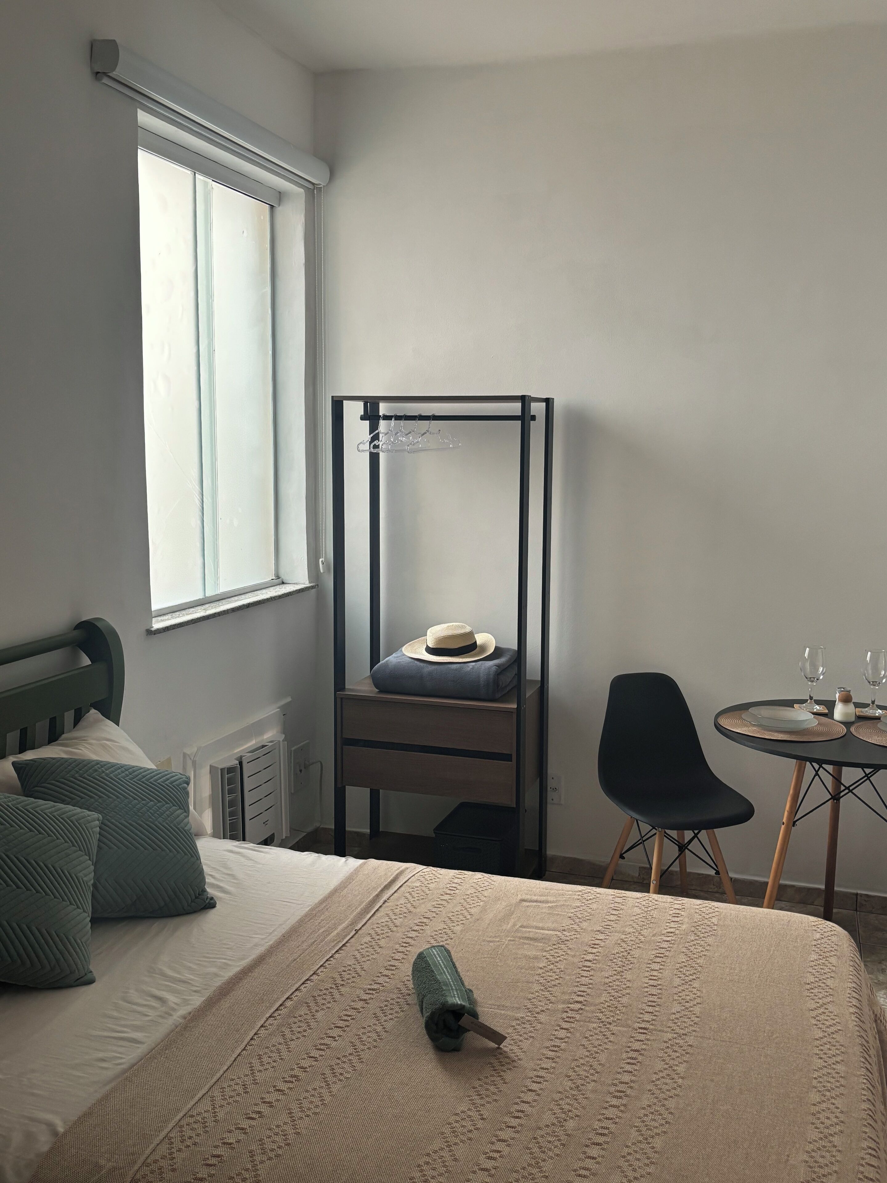 A bedroom with a bed, a small table with glasses, a chair, a window, and a metal shelf unit.