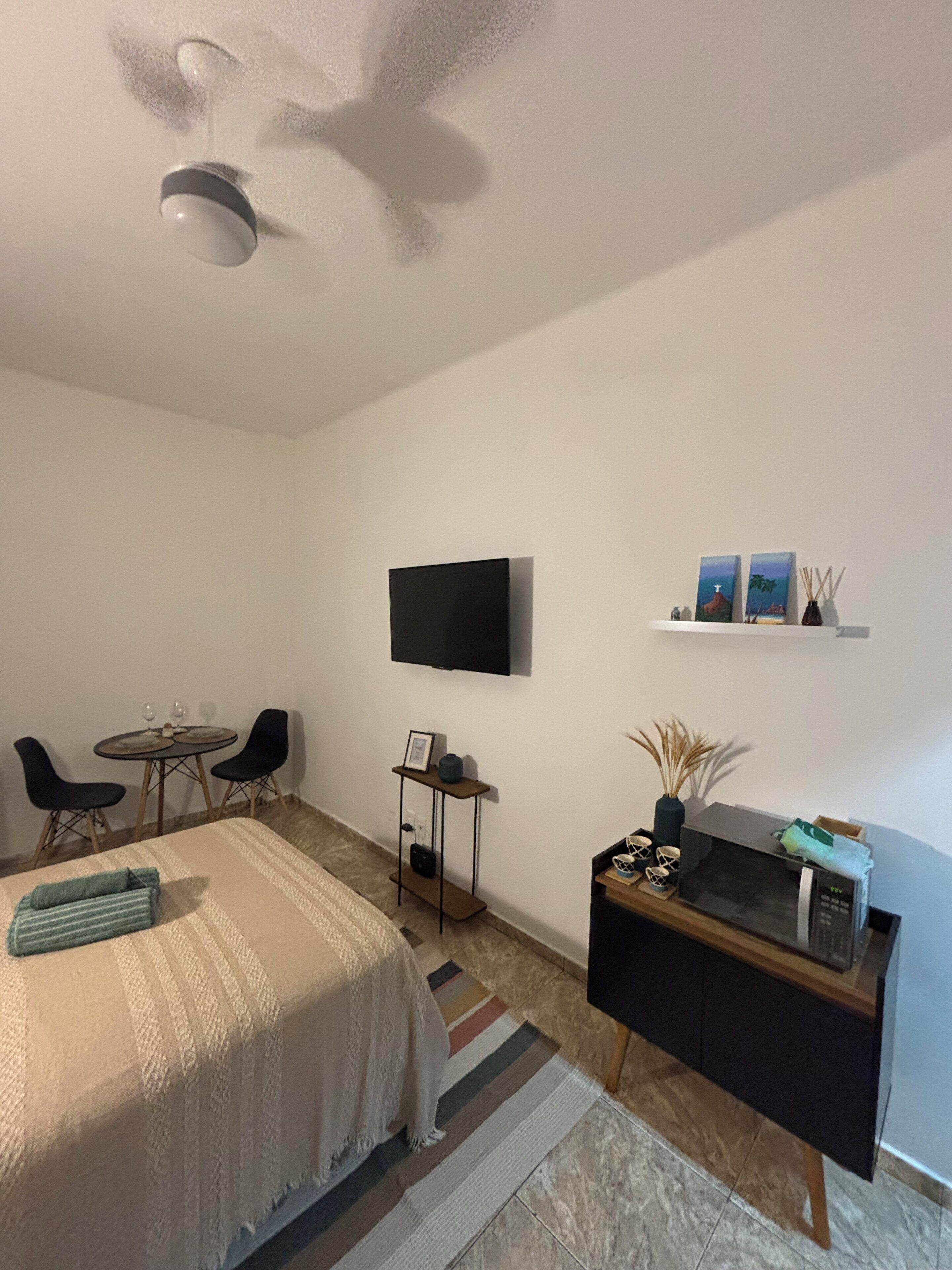 Une chambre d'hôtel de taille réduite comprenant un lit, un petit coin repas, une télévision, une étagère avec des objets décoratifs et un four à micro-ondes.