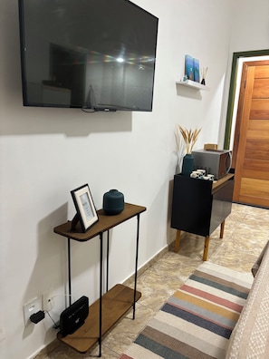 Ein modernes Wohnzimmer mit einem an der Wand befestigten Flachbildfernseher, einem Holztischchen mit Bilderrahmen und einer Vase, einem schwarzen Sessel und einem Streifenmuster-Teppich auf dem Boden.
