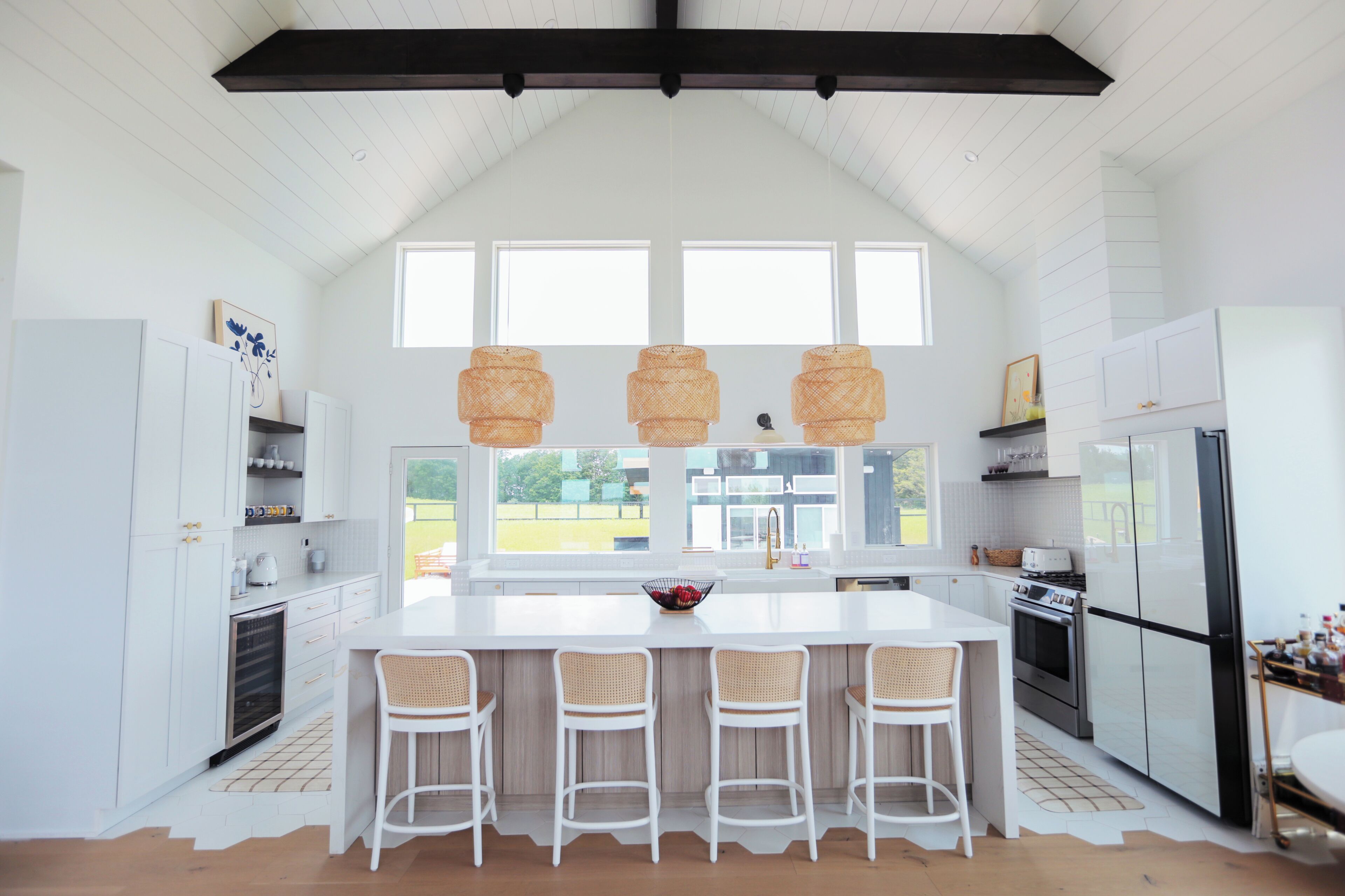 Una cocina moderna con armarios blancos, una isla grande con taburetes altos, lámparas colgantes y vistas a través de las ventanas.