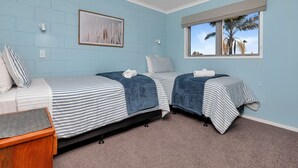 A bedroom with two beds, a window with a view, a framed picture on the wall, and a small wooden table. - The Lookout Waterfront Apartment at Harbour Lights (One Tree Point)