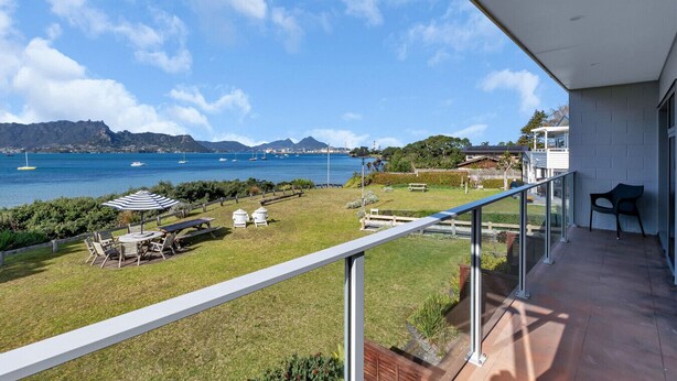 A balcony with a view of the sea, a grassy area with outdoor furniture, and a building with a white exterior. - The Lookout Waterfront Apartment at Harbour Lights (One Tree Point)