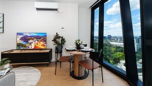 Ett modernt vardagsrum med en platt-TV, ett matbord med stolar, en krukväxt och ett stort fönster med utsikt över staden.