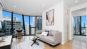 Una sala de estar moderna con sofá, mesa de centro y zona de comedor, ventanas amplias con vistas a la ciudad y un cuadro en la pared.