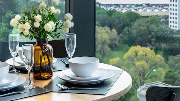 Mesa de comedor con un jarrón de flores, platos y vasos, con vistas a un paisaje urbano a través de un ventanal grande.