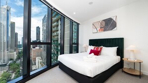 Una habitación de hotel moderna con una cama grande, vistas de la ciudad a través de ventanas de techo a suelo, un cabecero verde oscuro y una mesita de noche con una lámpara.