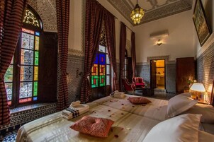 Una habitación con cama, una silla, una lámpara y una ventana con cortinas estampadas.