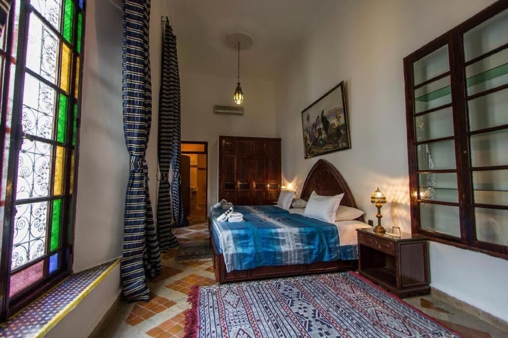 Un dormitorio con cama, mesita de noche, lámpara, ventana con cortinas y un cuadro en la pared.