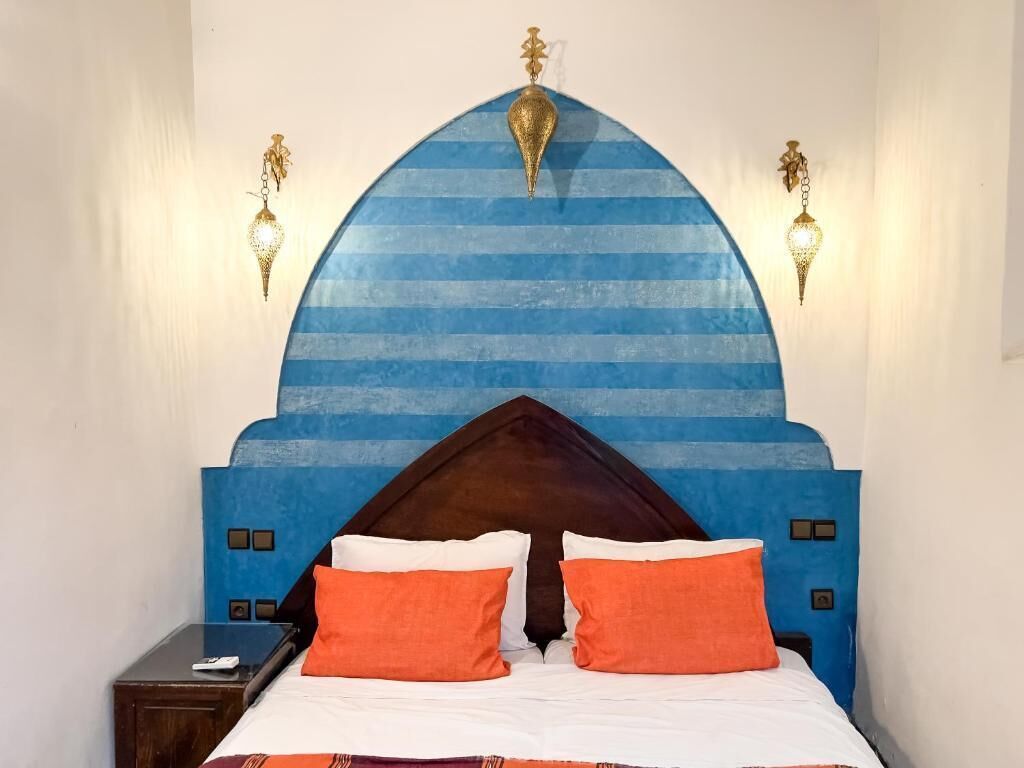 Un lit avec une tête de lit bleue, deux oreillers orange et un pied de lit en bois.