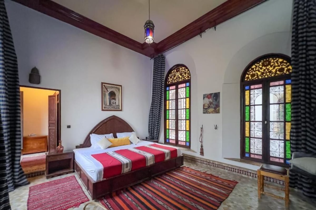 Una stanza con un letto grande, una testiera in legno, un comodino, una coperta a righe rosse e bianche, un tappeto decorato e uno sgabello di legno.