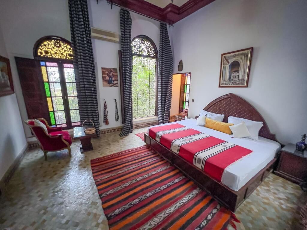 Une chambre avec un grand lit, une chaise, une petite table et une fenêtre avec des rideaux.