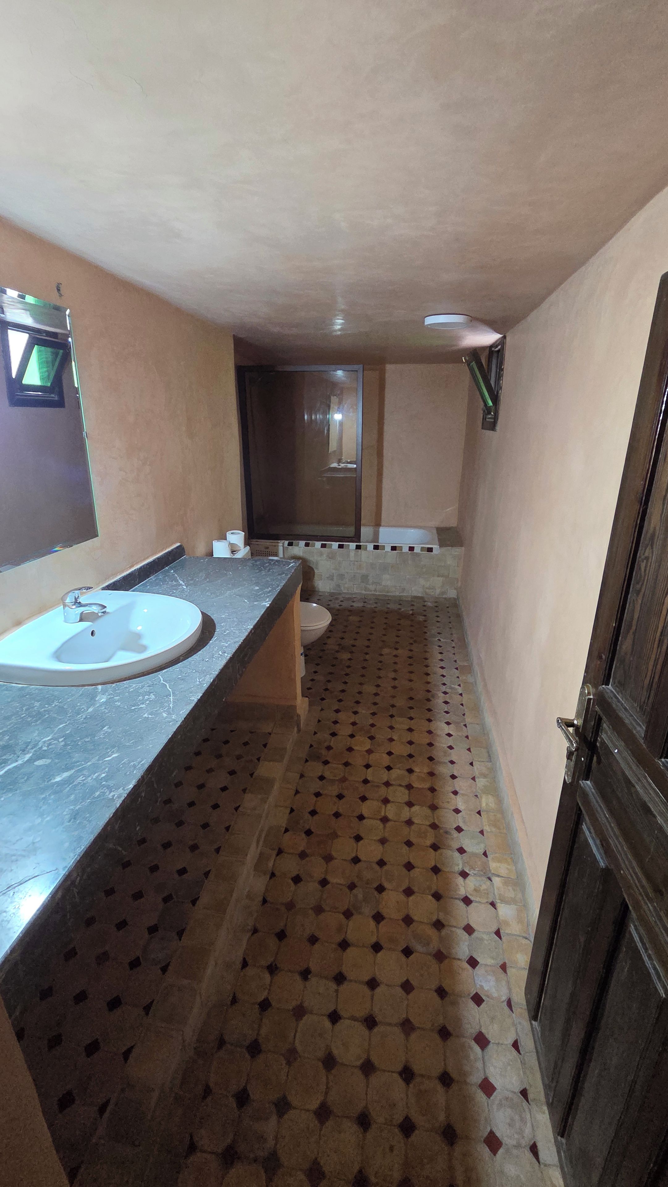 A narrow hallway with a sink and mirror on the left, a toilet and mirror on the right, and a patterned floor.