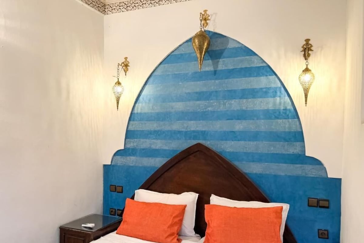 Camera da letto con una testiera blu, due cuscini arancioni, un comodino in legno e luci a parete.
