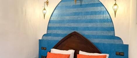 A bedroom with a blue headboard, two orange pillows, a wooden nightstand, and wall-mounted lights.