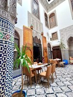 Un patio con suelo de baldosas, una zona de comedor con sillas y mesas, y detalles arquitectónicos ornamentales.