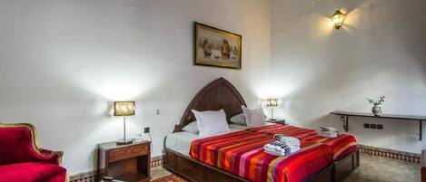 Une chambre d’hôtel avec un lit, un fauteuil rouge, une petite table, une table de chevet, une étagère fixée au mur et un tableau.