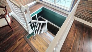 Una escalera con barandilla blanca que baja a un nivel inferior, una ventana con vistas al exterior y una pared de ladrillo.