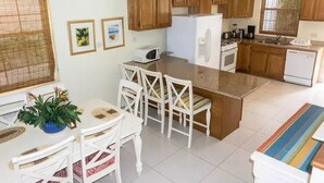 A kitchen with wooden cabinets, a white refrigerator, a microwave, and a dining area with white chairs and a table. - Unit 6 Coast Luxury Town Homes  Newly Refurbished (Rodney Bay)