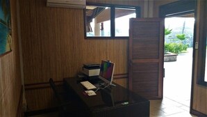 Een kamer met een houten wand, een bureau met een computer, een stoel en een raam met uitzicht op groen.