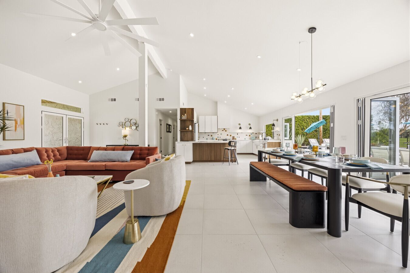 A spacious living area with a sofa, a dining table with chairs, a kitchen in the background, and large windows.