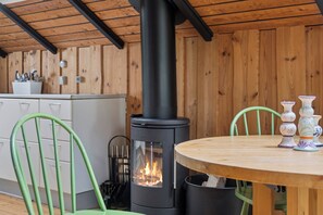 A room with a wood-burning stove, green chairs, a wooden table, and a kitchen area with cabinets and utensils.