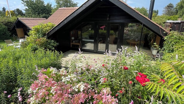 Een houten blokhut met een schuin dak, grote glasramen en een tuin met verschillende bloemen.