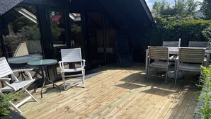 Een houten terras met buitenmeubilair, waaronder tafels en stoelen, en een glazen deuropening.
