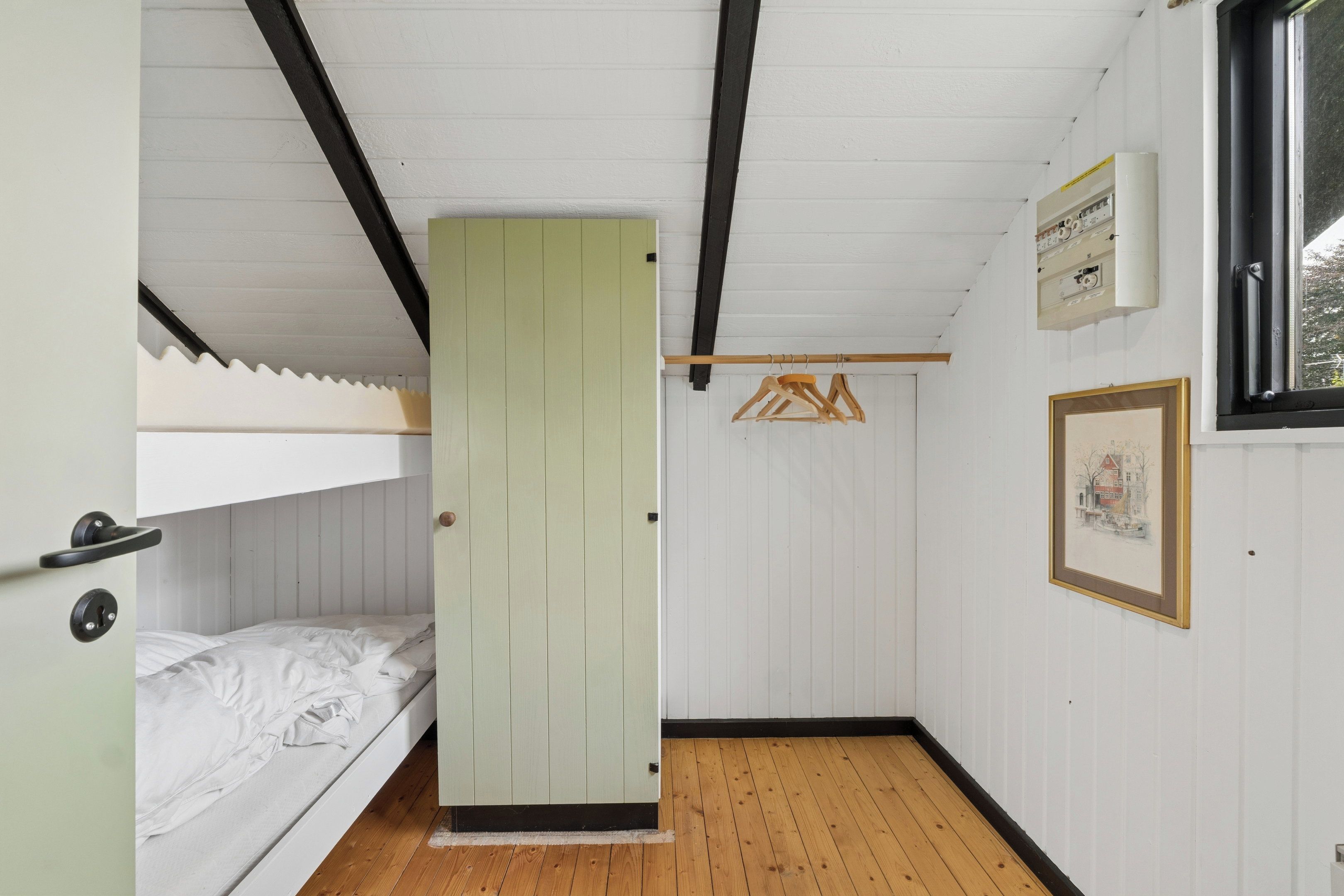 Un petit logement composé d’une seule pièce, avec un lit superposé, un plancher en bois, une porte, un porte-manteau et un tableau encadré au mur.