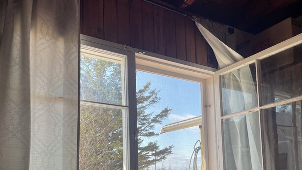 A room with wooden walls, a large window with curtains, and a broom leaning against the wall. - South shore time capsule (Canoe Cove)