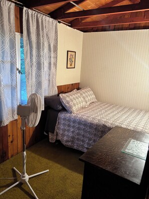 A small, cozy room with a single bed, a standing fan, and a wooden table. - South shore time capsule (Canoe Cove)