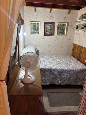 A room with a bed, a standing fan, and two framed paintings on the wall.