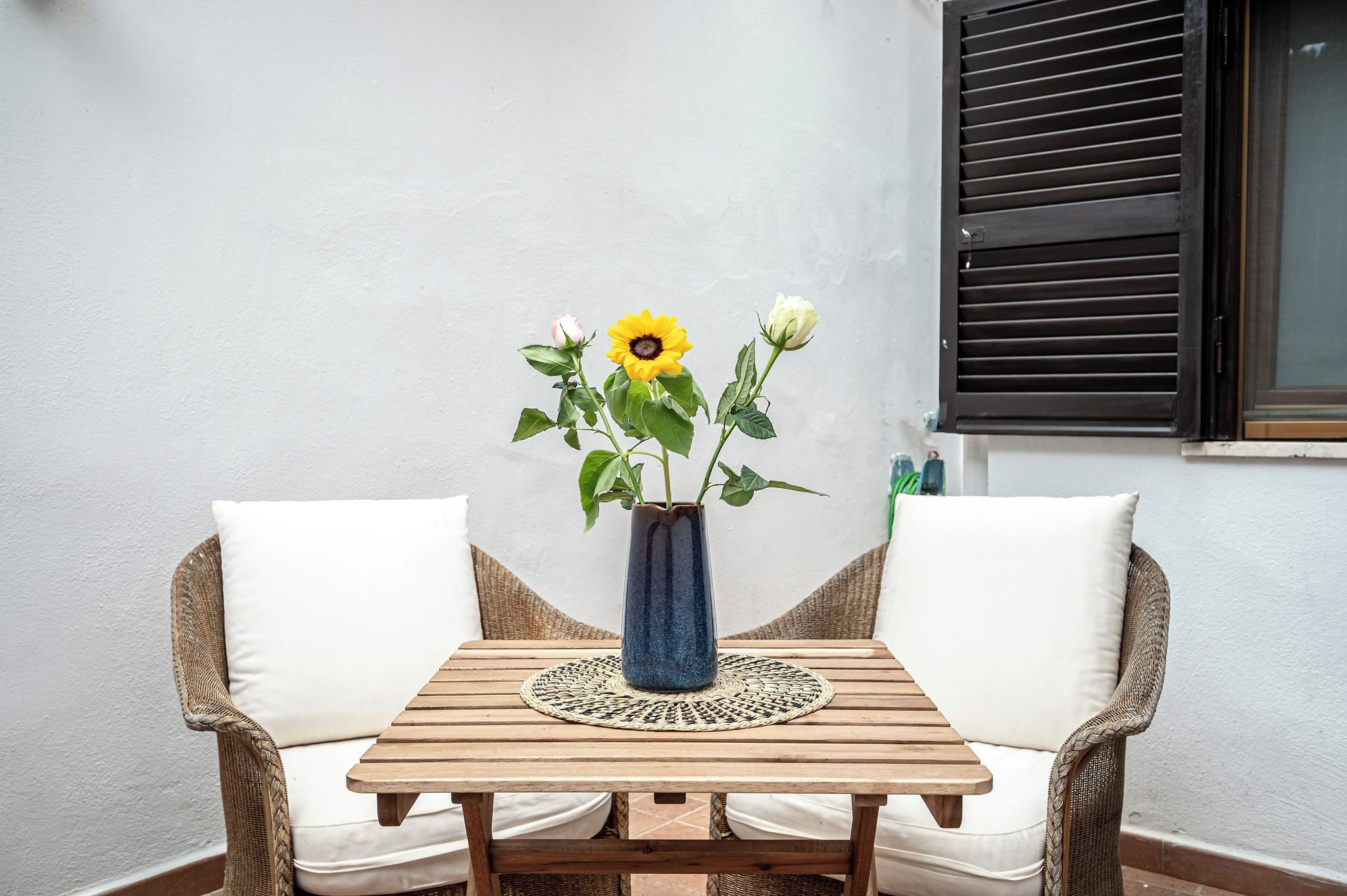 A wooden table with a vase of flowers, two wicker chairs, and a window with blinds.