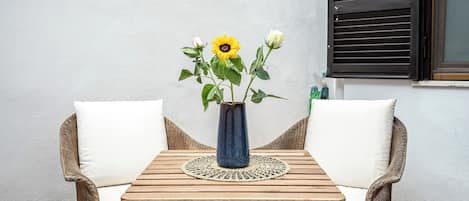 A wooden table with a vase of flowers, two wicker chairs, and a window with blinds.