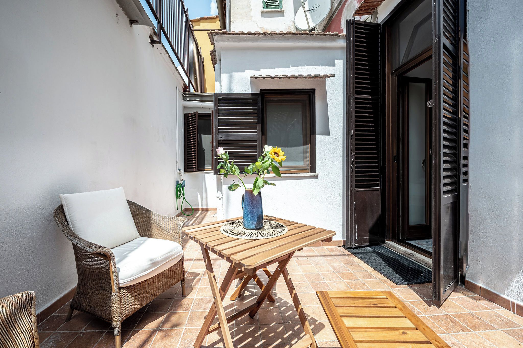 A small outdoor seating area with a wicker chair, a wooden table, and a vase with flowers.