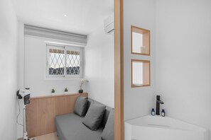 A modern room with a grey sofa, a small window with a view, a white sink, and a mirror.
