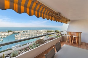 Ein Balkon mit Blick auf einen Yachthafen und das Meer, ein Tisch mit zwei Stühlen und ein gestreifter Sonnenschirm.